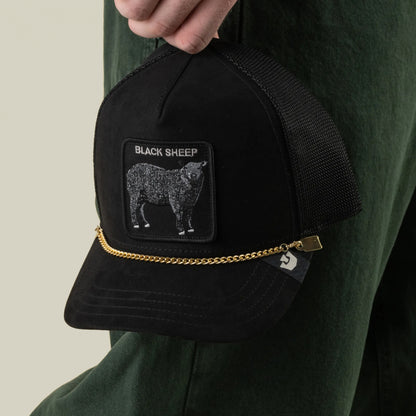 A hand holds a black mesh trucker hat by Goorin Bros., featuring a BLACK SHEEP patch, a sheep graphic, and the stylish Mini CapLoc Chain—hat jewelry with a gold chain and double clip chains adorning the front. The person is wearing dark green pants.