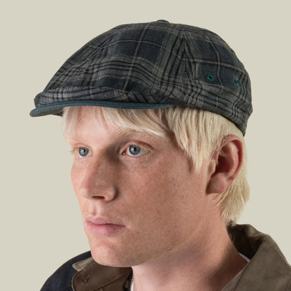 A person with blonde hair wears the Goorin Bros. Point Lobos gray plaid 6 Panel Ivy flat cap, handmade in the USA from a breathable cotton blend, along with a brown collared jacket, shown against a plain light background.