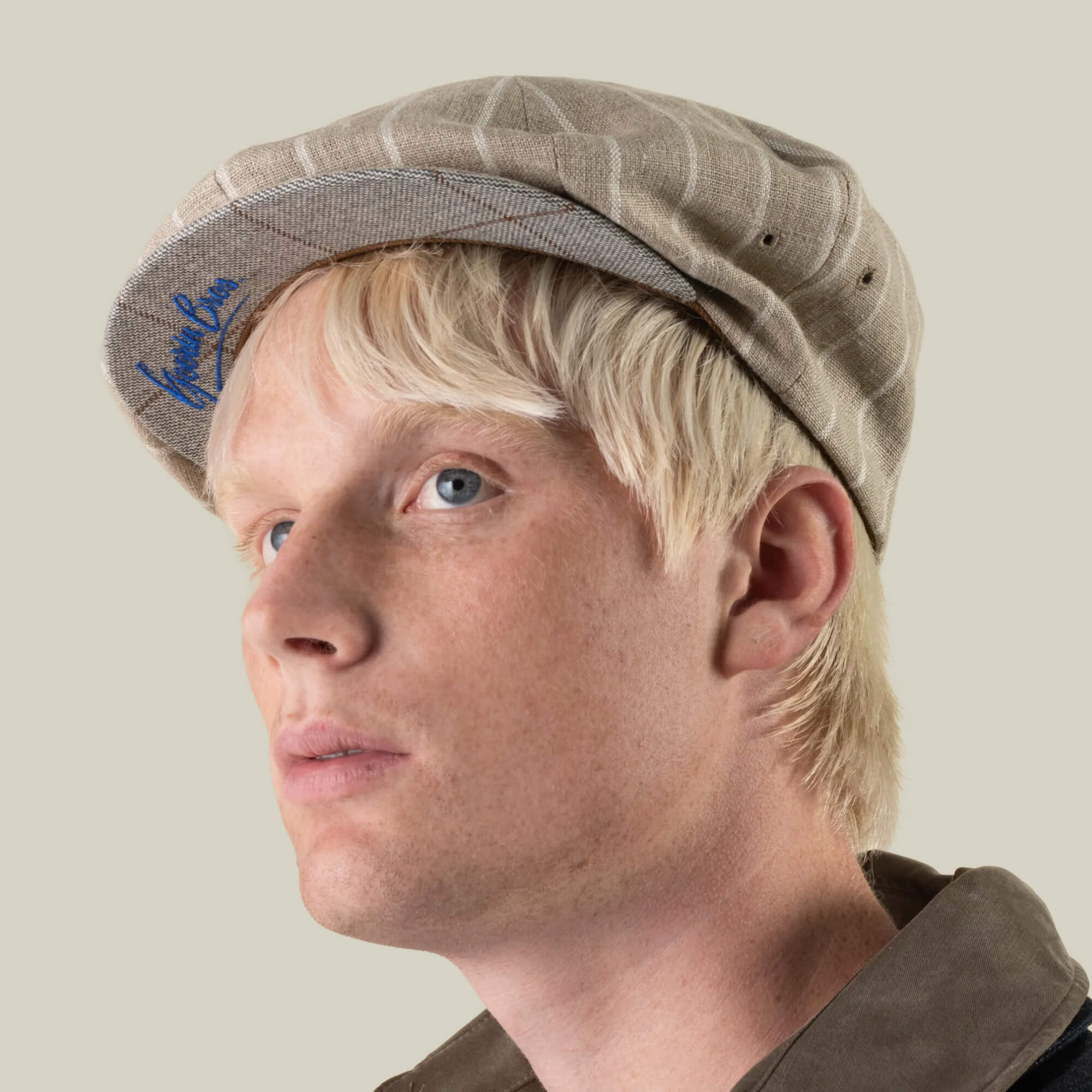 A person with light blond hair wears the Rincon Park cap by Goorin Bros.—a beige plaid classic Gatsby silhouette—paired with a brown shirt, looking slightly to the side against a plain background. The hand cut and sewn construction from a soft linen blend fabric lends an artisanal touch to this timeless piece.