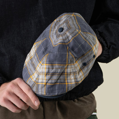 A person holds the Goorin Bros. Point Lobos 6 Panel Ivy flat cap—featuring a blue and yellow plaid pattern—against their side while wearing a dark long-sleeve top and olive pants; this cap is handmade in the USA from a breathable cotton blend.