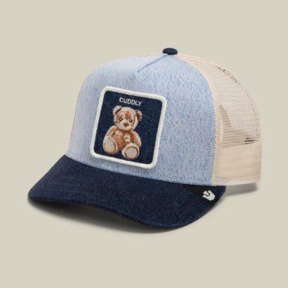 The Goorin Bros. Denim Cuddly trucker hat features a light blue denim front, navy blue bill, cream mesh back panels, and an embroidered teddy bear patch on the crown, shown against a neutral studio background.