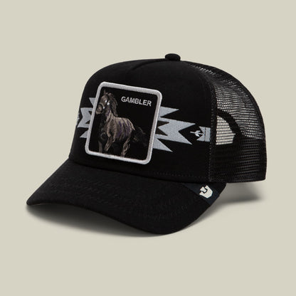 The Canyon Gambler by Goorin Bros is a black trucker hat with a mesh back, featuring an embroidered rooster patch labeled "Gambler" and white lightning bolt designs on the front.